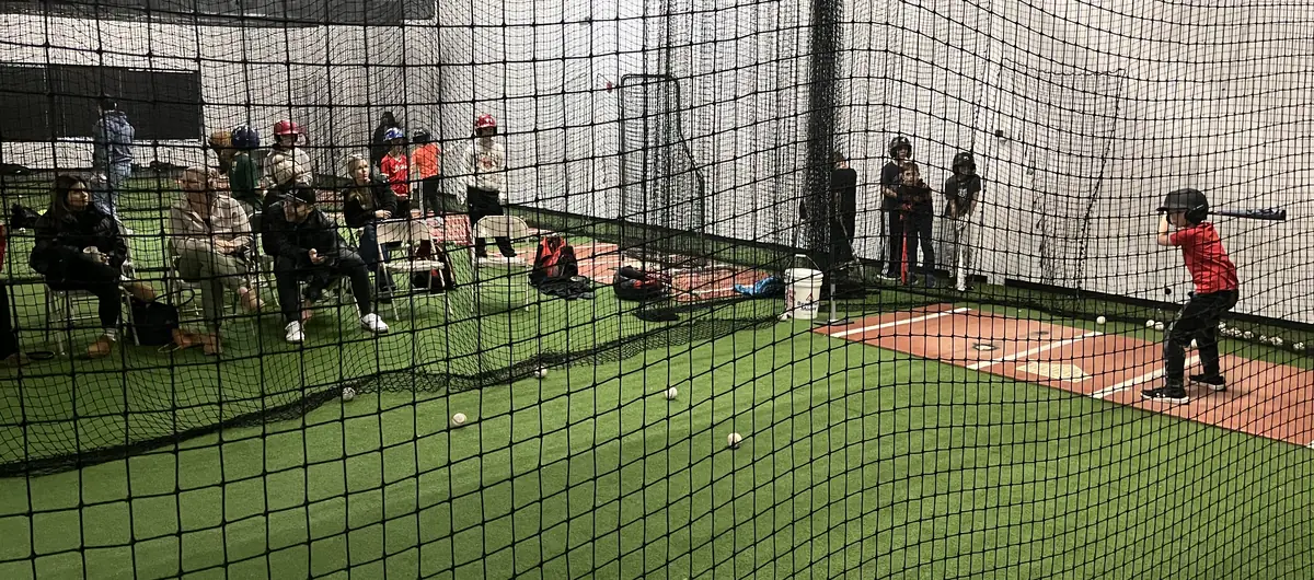 Parents watching from chairs beside the cage as a young player waits for the pitch